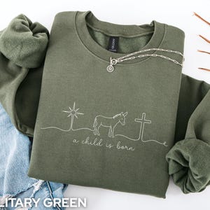 May include: A military green crewneck sweatshirt featuring a white line art graphic of a star, a donkey, and a cross, with the text "a child is born" written in a cursive script below the design. The sweatshirt has long sleeves and a fleece lining.