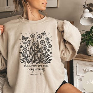 Puede incluir: Sudadera color arena con un gráfico negro de dientes de león y mariposas. El texto "His mercies are new every morning, Lamentations 3:23" está impreso debajo. La sudadera tiene cuello redondo y mangas largas.