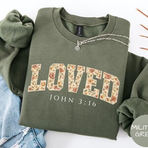 Puede incluir: Una sudadera verde oliva con la palabra "LOVED" en un diseño floral en la parte delantera. Debajo está el texto "JOHN 3:16". Se ve un collar plateado con un colgante circular.