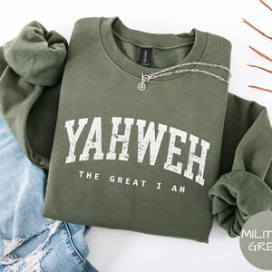 Puede incluir: Sudadera verde militar con el texto blanco "YAHWEH THE GREAT I AM", combinada con vaqueros azules y un collar plateado. La sudadera tiene cuello redondo y puños acanalados.