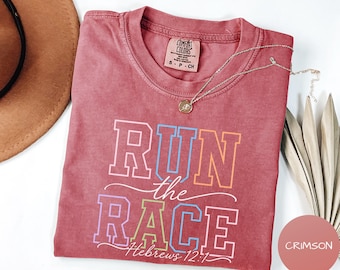 May include: A crimson short-sleeve t-shirt with a crew neckline and a Comfort Colors label. The front features the text RUN THE RACE and Hebrews 12:1 in a multi-colored block and script font. A thin gold-tone chain necklace rests on the fabric.