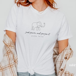 May include: White t-shirt featuring a minimalist line drawing of an elephant and the text "seek peace, and pursue it psalm 34:14" in black script. The shirt is made of a soft material and has a classic crew neck.