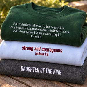 May include: A stack of three folded crewneck sweatshirts. The top is dark green with white text reading John 3:16. The middle is white with red text reading strong and courageous Joshua 1:9. The bottom is charcoal gray with white text reading DAUGHTER OF THE KING.
