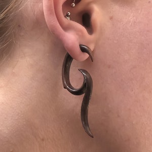 May include: A person's ear with multiple piercings. The ear is pierced with a helix piercing, a conch piercing, and a lobe piercing. The lobe piercing has a large, dark brown, wooden, curved earring.