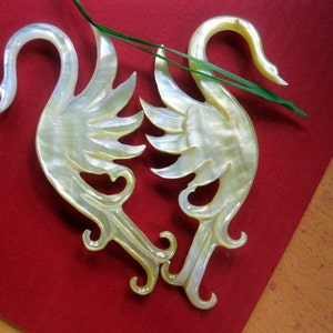 May include: Two white mother of pearl swan ornaments with intricate details. The swans are facing each other with their necks curved.