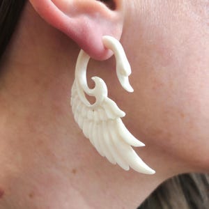 May include: A pair of white bone earrings shaped like wings with intricate carvings. The earrings are large and dangle from the earlobe.