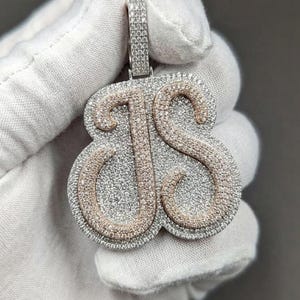 May include: A silver-toned pendant with the letters "JS" encrusted with small, sparkling stones. The letters are outlined in a rose gold color, and the pendant is held by a white-gloved hand.