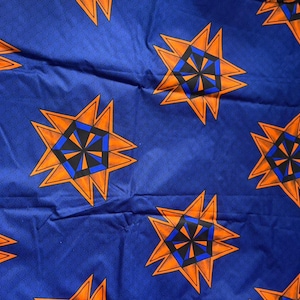 Op de afbeelding: Een blauwe stof met een herhalend geometrisch patroon van oranje, zwarte en blauwe stervormige figuren. Elke ster bestaat uit een oranje buitenrand met een centraal zwart en blauw zeshoekig ontwerp, wat zorgt voor een levendige grafische print.