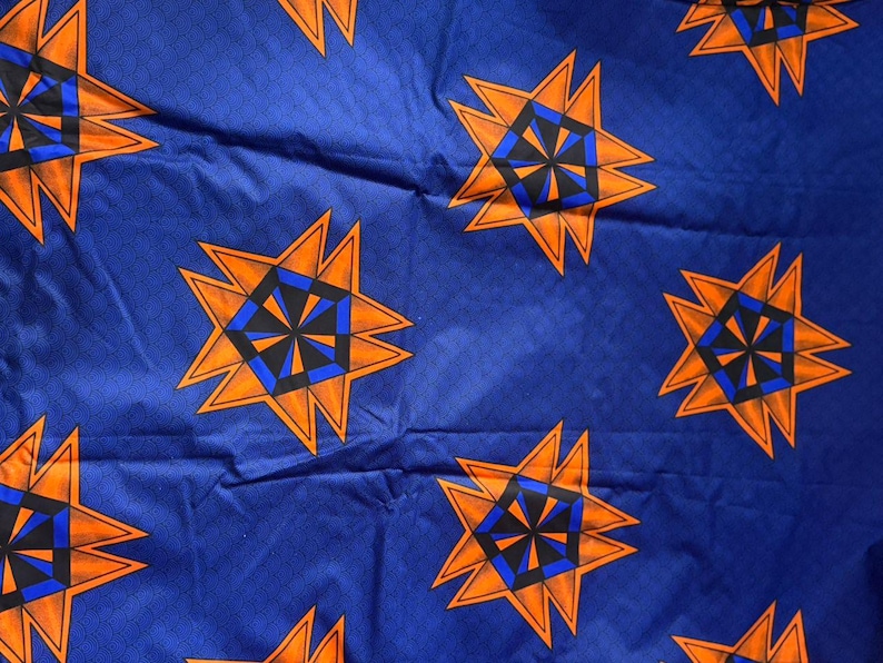 Op de afbeelding: Een blauwe stof met een herhalend geometrisch patroon van oranje, zwarte en blauwe stervormige figuren. Elke ster bestaat uit een oranje buitenrand met een centraal zwart en blauw zeshoekig ontwerp, wat zorgt voor een levendige grafische print.
