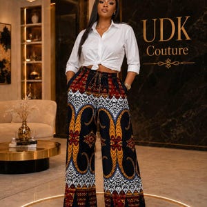 May include: A white button-down shirt with long sleeves tied at the waist, paired with wide-leg trousers featuring a black base and a repeating geometric pattern in shades of red, yellow, and white. The trousers have a drawstring waist.