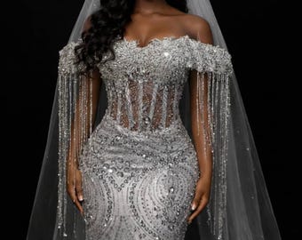 Op de afbeelding: Een zilveren off-the-shoulder trouwjurk met ingewikkeld kralenwerk en een vloeiend zeemeerminnen silhouet. De jurk heeft een doorschijnend lijfje en kralenfranjes op de mouwen. Een doorschijnende sluier en een versierd diadeem maken de look compleet.