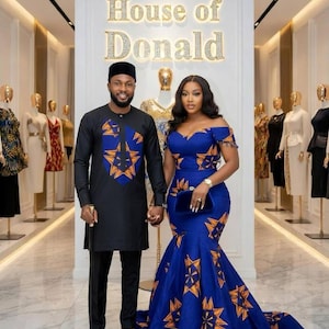 Op de afbeelding: Een zwarte tuniek met lange mouwen met een blauw en oranje sterpatroon op de borst, een zwarte broek en een zwarte pet, naast een blauwe off-shoulder zeemeermin-jurk met een oranje sterpatroon, staand voor een House of Donald bord.