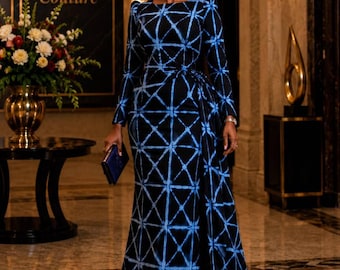 Op de afbeelding: Een vloerlange jurk met lange mouwen in zwart met een blauw ruitpatroon. Het kledingstuk heeft een aansluitend lijfje, een hoge halslijn en een gedrapeerd zijpaneel dat tot op de grond reikt. De stof vertoont een tie-dye effect in de lijnen.