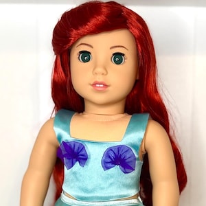 May include: A doll with red hair and green eyes, wearing a light blue satin top and skirt. The top has purple accents. The doll is standing and facing the camera.