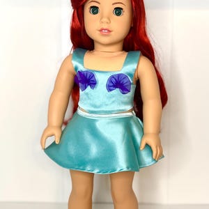 May include: A doll with red hair and green eyes, wearing a light blue satin top and skirt. The top has purple accents. The doll is standing and facing the camera.