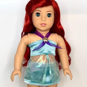K&ouml;nnte beinhalten: Puppe mit rotem Haar und gr&uuml;nen Augen, bekleidet mit einem hellblauen Oberteil mit Muschelakzent und einem passenden Rock. Das Outfit der Puppe hat einen schimmernden, irisierenden &Uuml;berzug. Die Puppe ist eine beliebte Figur.