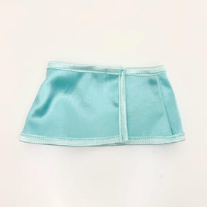 Op de afbeelding: Een kleine, lichtblauwe satijnen rok met een rechte zoom. De rok heeft een naad aan de achterkant en is afgezet met een lichter blauw satijnen lint langs de zoom en tailleband. De rok is ongeveer 15 cm breed.