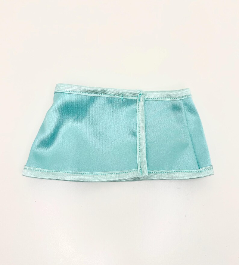 Op de afbeelding: Een kleine, lichtblauwe satijnen rok met een rechte zoom. De rok heeft een naad aan de achterkant en is afgezet met een lichter blauw satijnen lint langs de zoom en tailleband. De rok is ongeveer 15 cm breed.