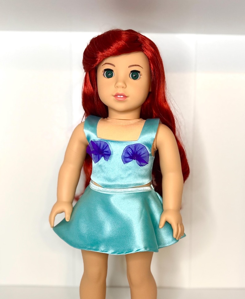 May include: A doll with red hair and green eyes, wearing a light blue satin top and skirt. The top has purple accents. The doll is standing and facing the camera.