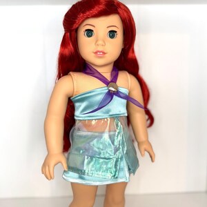 K&ouml;nnte beinhalten: Puppe mit rotem Haar und gr&uuml;nen Augen, bekleidet mit einem hellblauen Satin-Oberteil und einem passenden, schillernden Rock. Ein lila Band ist um den Hals gebunden, und ein muschelf&ouml;rmiger Verschluss ziert das Oberteil. Ein beliebtes Kinderspielzeug.