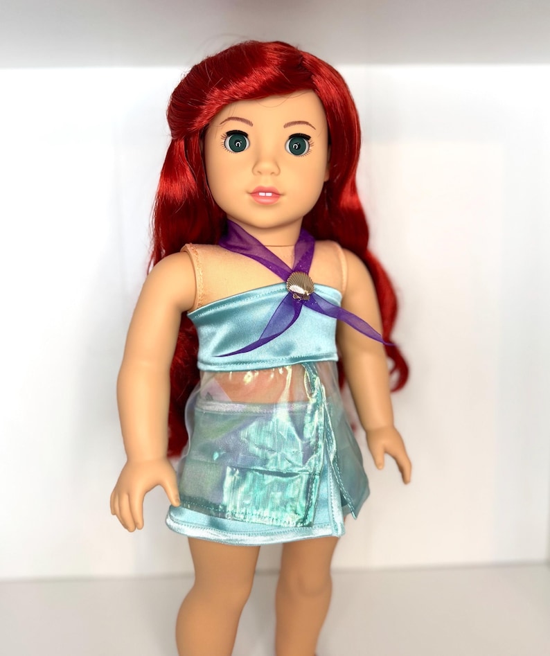 K&ouml;nnte beinhalten: Puppe mit rotem Haar und gr&uuml;nen Augen, bekleidet mit einem hellblauen Satin-Oberteil und einem passenden, schillernden Rock. Ein lila Band ist um den Hals gebunden, und ein muschelf&ouml;rmiger Verschluss ziert das Oberteil. Ein beliebtes Kinderspielzeug.