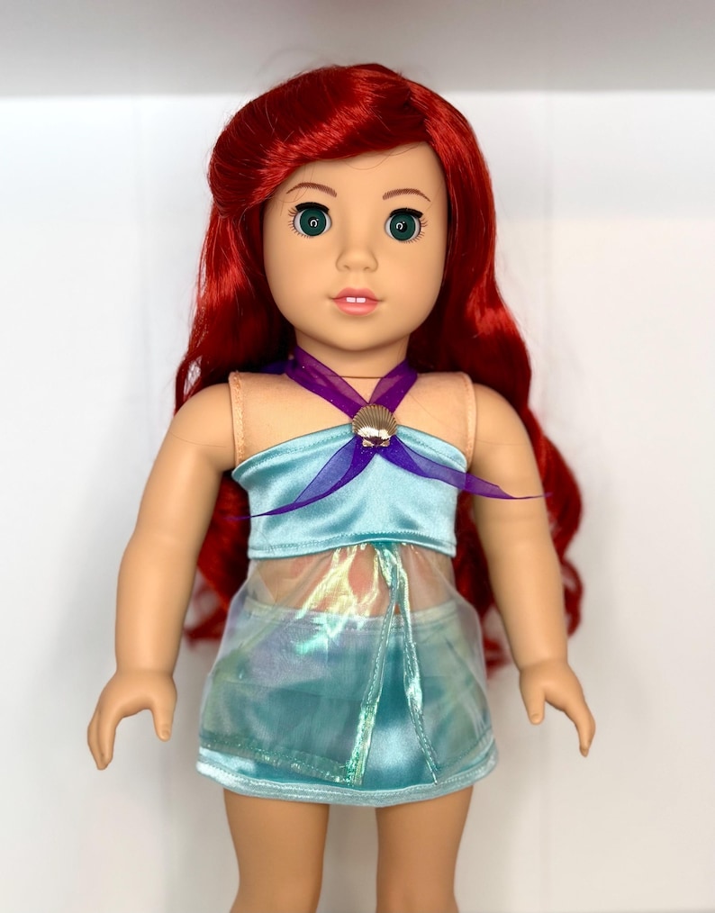 K&ouml;nnte beinhalten: Puppe mit rotem Haar und gr&uuml;nen Augen, bekleidet mit einem hellblauen Oberteil mit Muschelakzent und einem passenden Rock. Das Outfit der Puppe hat einen schimmernden, irisierenden &Uuml;berzug. Die Puppe ist eine beliebte Figur.