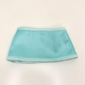Op de afbeelding: Een kleine, turquoise rok gemaakt van een glanzende stof. De rok heeft een rechte snit en is aan de boven- en onderkant afgezet met een lichtere blauwe stof. Waarschijnlijk ontworpen voor een pop of een kleine figuur.
