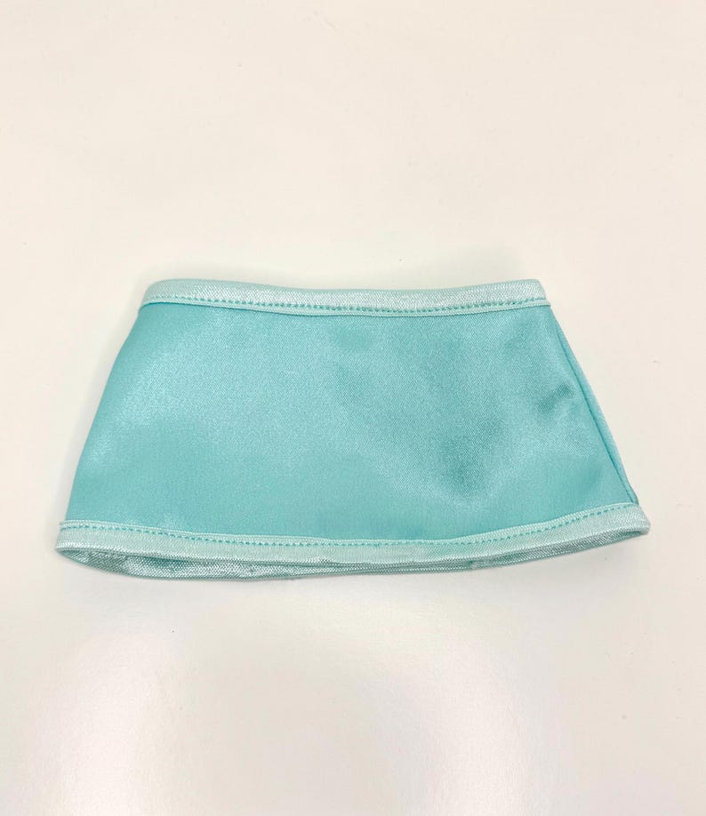 Op de afbeelding: Een kleine, turquoise rok gemaakt van een glanzende stof. De rok heeft een rechte snit en is aan de boven- en onderkant afgezet met een lichtere blauwe stof. Waarschijnlijk ontworpen voor een pop of een kleine figuur.