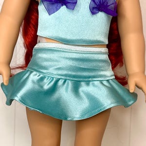 Op de afbeelding: Een pop met een lichtblauwe satijnen tweedelige outfit. De top heeft twee paarse stoffen accenten en de rok is gerimpeld. De pop heeft rood haar. De outfit is ontworpen om op een zeemeerminkostuum te lijken.