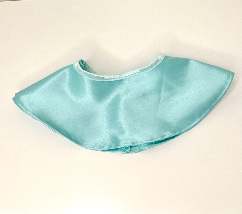 May include: A light blue satin cape with a rounded neckline. The cape is a solid color and has a smooth, shiny texture. The cape is designed to be worn around the neck and shoulders.