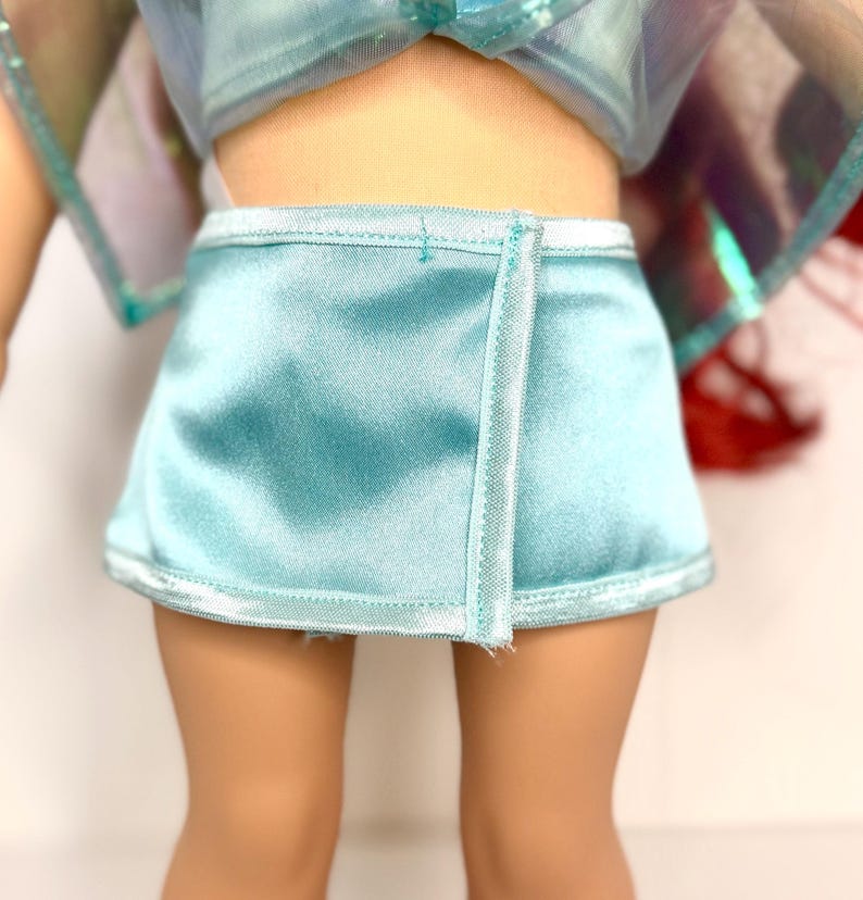 Op de afbeelding: Een lichtblauwe satijnen wikkelrok met witte bies. De rok maakt deel uit van een poppenoutfit en heeft een eenvoudig, elegant ontwerp. De rok is ongeveer 10 cm lang.
