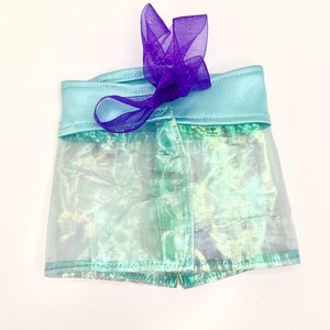 K&ouml;nnte beinhalten: Schimmernde Shorts mit hellblauem Bund und einem transparenten, aquafarbenen Overlay. Eine lila, glitzernde Schleife ziert die Vorderseite. Die Shorts bestehen aus leichtem Material. Die Shorts sind etwa 20 cm lang.