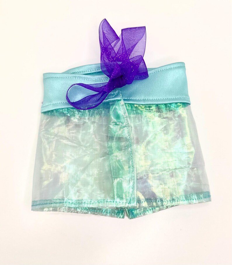 K&ouml;nnte beinhalten: Schimmernde Shorts mit hellblauem Bund und einem transparenten, aquafarbenen Overlay. Eine lila, glitzernde Schleife ziert die Vorderseite. Die Shorts bestehen aus leichtem Material. Die Shorts sind etwa 20 cm lang.