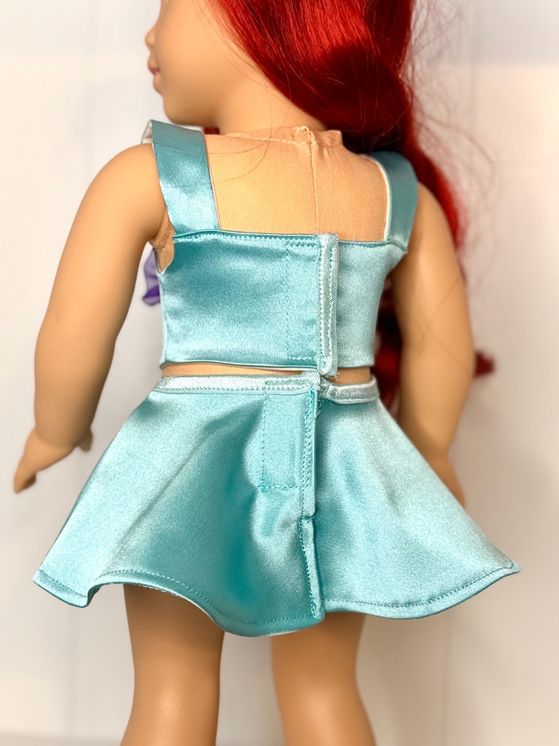 May include: A doll wearing a light blue satin two-piece outfit. The top has square straps and a zipper closure in the back. The skirt is a matching light blue satin material.