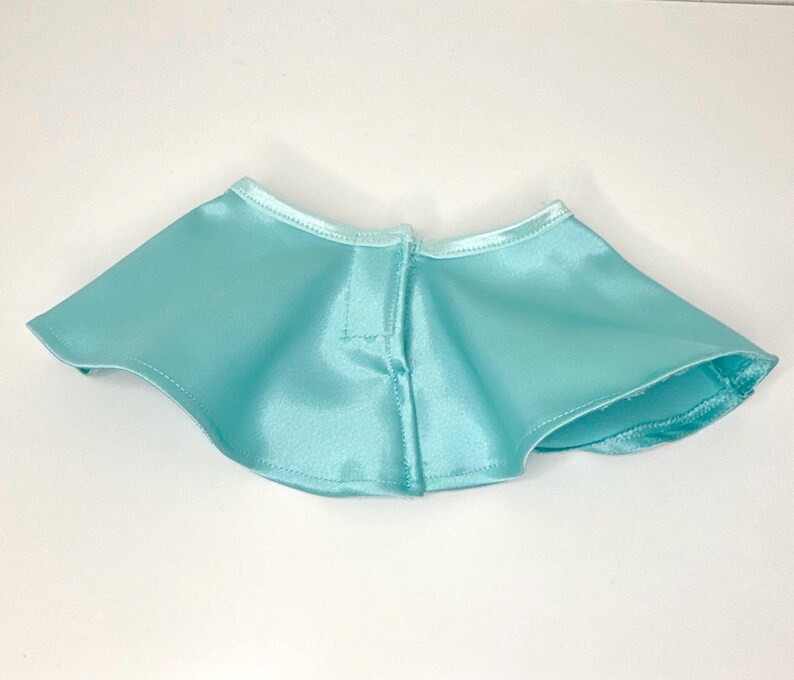 May include: A light blue satin cape with a rounded shape. The cape has a small collar and a button closure at the front. The fabric has a slight sheen, and the edges are neatly finished with a matching trim.