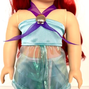 K&ouml;nnte beinhalten: Eine Puppe tr&auml;gt ein hellblaues Satin-Top mit einem lila Tr&auml;gerband und einem goldenen Muscheldetail. Die Puppe tr&auml;gt auch einen durchsichtigen, schillernden Rock. Die Puppe hat rotes Haar.