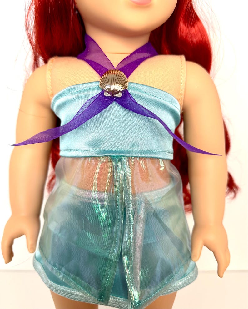 K&ouml;nnte beinhalten: Eine Puppe tr&auml;gt ein hellblaues Satin-Top mit einem lila Tr&auml;gerband und einem goldenen Muscheldetail. Die Puppe tr&auml;gt auch einen durchsichtigen, schillernden Rock. Die Puppe hat rotes Haar.