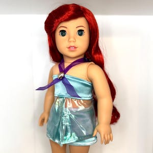 Op de afbeelding: Pop met rood haar en groene ogen, gekleed in een lichtblauwe top en rok. De top heeft een paars lint en een schelpvormige versiering. De rok heeft een glanzende, iriserende uitstraling. Een populair personage.