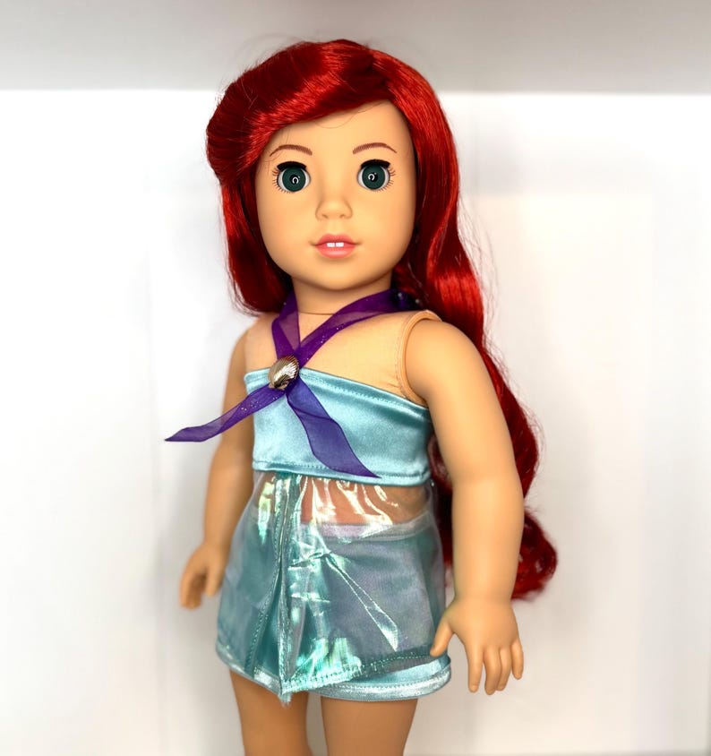 Op de afbeelding: Pop met rood haar en groene ogen, gekleed in een lichtblauwe top en rok. De top heeft een paars lint en een schelpvormige versiering. De rok heeft een glanzende, iriserende uitstraling. Een populair personage.