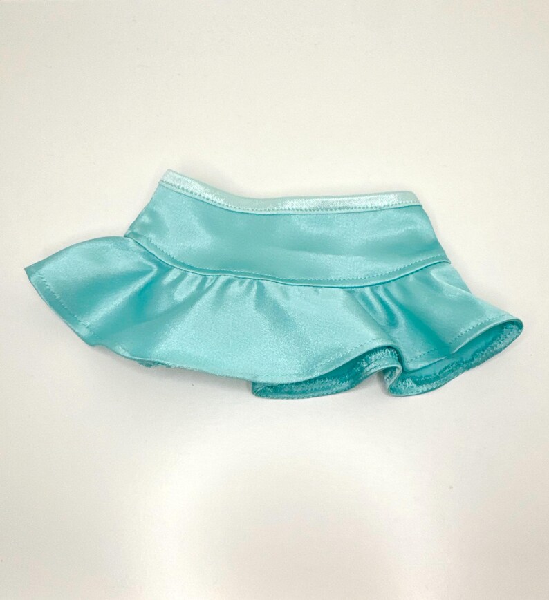 Op de afbeelding: Een lichtblauwe satijnen rok met een gefranjerde zoom. De rok heeft een gladde, glanzende textuur en een getailleerde tailleband. De rok is ontworpen voor een pop of een ander klein figuur.