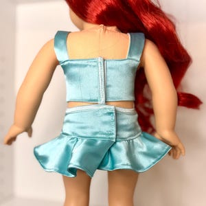 Op de afbeelding: Pop in een lichtblauwe satijnen tweedelige outfit. De top is een crop top met vierkante bandjes en de rok is een minirok met een geplooid zoom. De pop heeft lang rood haar.