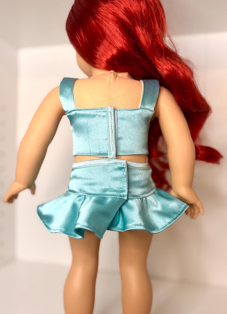 Op de afbeelding: Pop in een lichtblauwe satijnen tweedelige outfit. De top is een crop top met vierkante bandjes en de rok is een minirok met een geplooid zoom. De pop heeft lang rood haar.