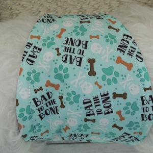 May include: A light blue dog bandana with a repeating pattern of brown bones, white skulls, and green paw prints. The phrase "BAD TO THE BONE" is printed in black and brown.