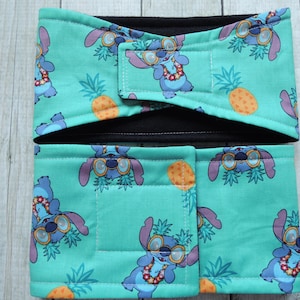 Puede incluir: Conjunto de dos piezas con tela turquesa con un estampado repetido de un personaje de dibujos animados azul con gafas de sol y un collar de flores, junto con piñas. El conjunto incluye una diadema y una pieza rectangular con cierre de velcro.