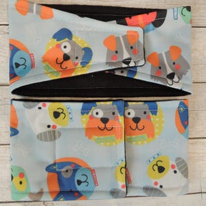 May include: Two blue and white fabric dog-themed headbands with black fleece lining. The fabric features a variety of cartoon dogs with different expressions and colors.