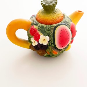 May include: A colorful ceramic teapot with a fruit design. The teapot features a pineapple lid, a watermelon slice, strawberries, and other fruits. The handle and spout are yellow, and the body is decorated with green leaves and flowers.