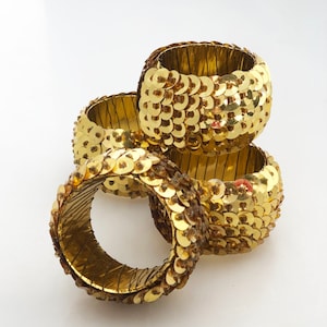 May include: Four gold sequin napkin rings. The rings are made of a wire base covered in gold sequins.