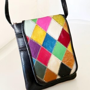 May include: A black crossbody bag featuring a flap with a multi-colored diamond patchwork pattern. The bag includes two visible zippered compartments on the side and a thin black shoulder strap. The patchwork consists of various colors including red, yellow, blue, green, purple, white, and brown leather-like segments.