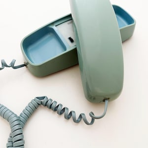 May include: A sage green landline telephone with a matching coiled cord. The handset is resting on the base unit, which features a light blue interior compartment and a small rectangular white panel with a black switch visible on the surface.