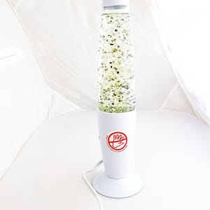 May include: A white lava lamp with a conical top and base. The clear glass cylinder contains green and silver glitter suspended in a clear liquid. The base has a red and white warning label. A white power cord extends from the base.